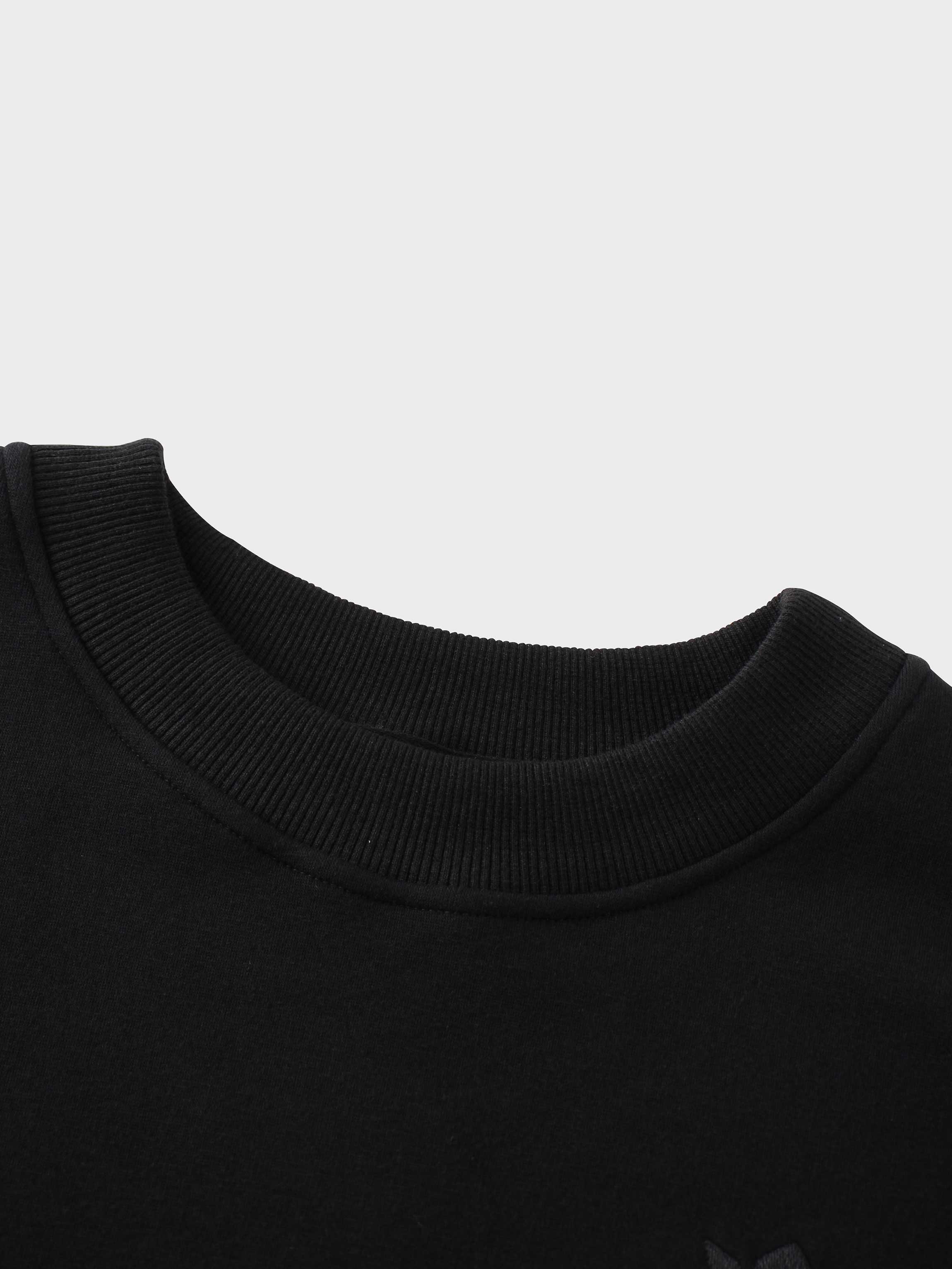 A close-up of the ribbed crewneck collar of the black sweatshirt. The top of the subtle, black-on-black embroidered logo is faintly visible on the chest area below the neckline.