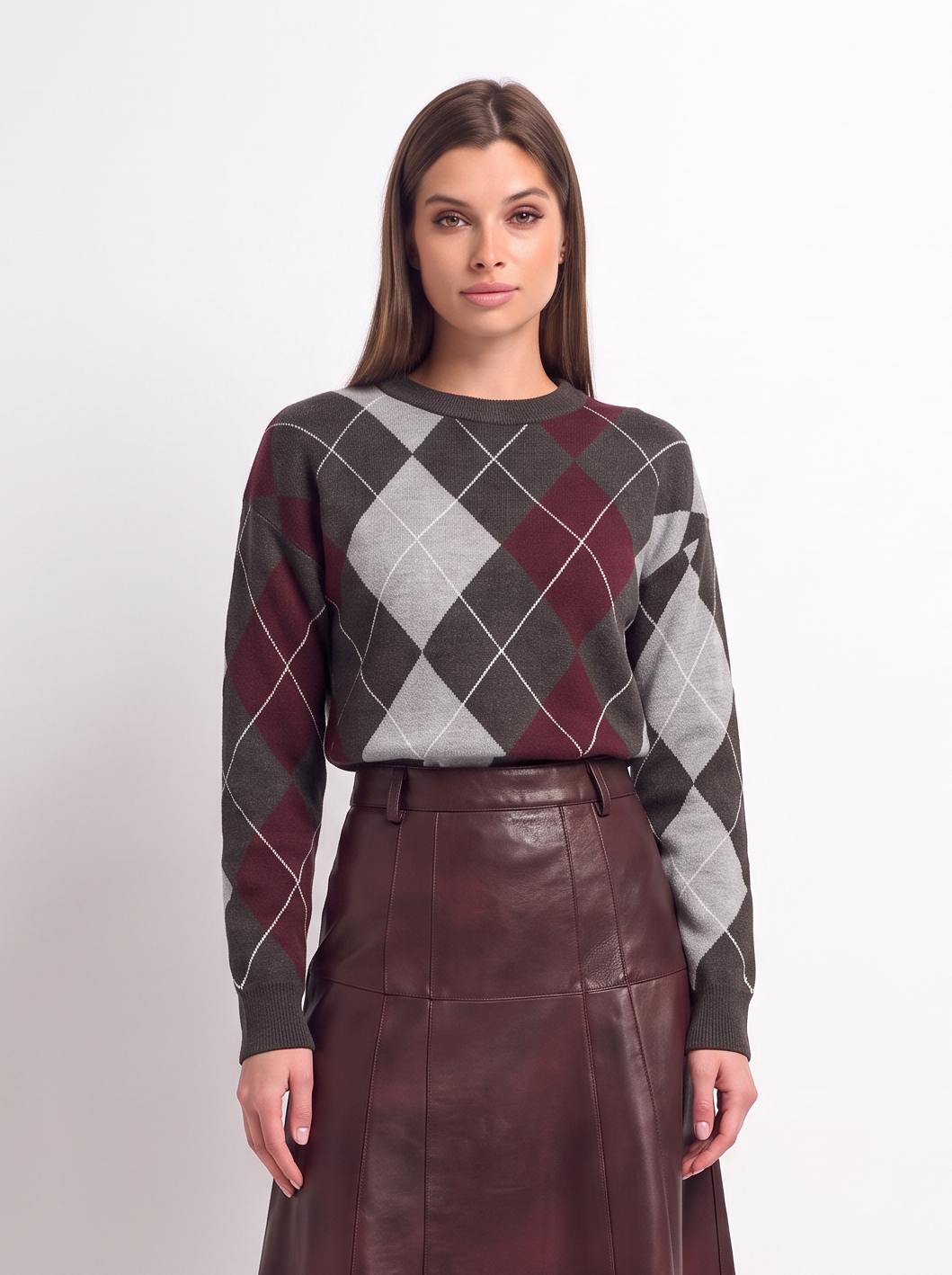 A woman stands against a plain light gray studio background wearing a long-sleeve argyle sweater in dark gray, burgundy, and white, paired with a dark burgundy pleated midi skirt and black pointed heels.