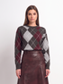 A woman stands against a plain light gray studio background wearing a long-sleeve argyle sweater in dark gray, burgundy, and white, paired with a dark burgundy pleated midi skirt and black pointed heels.
