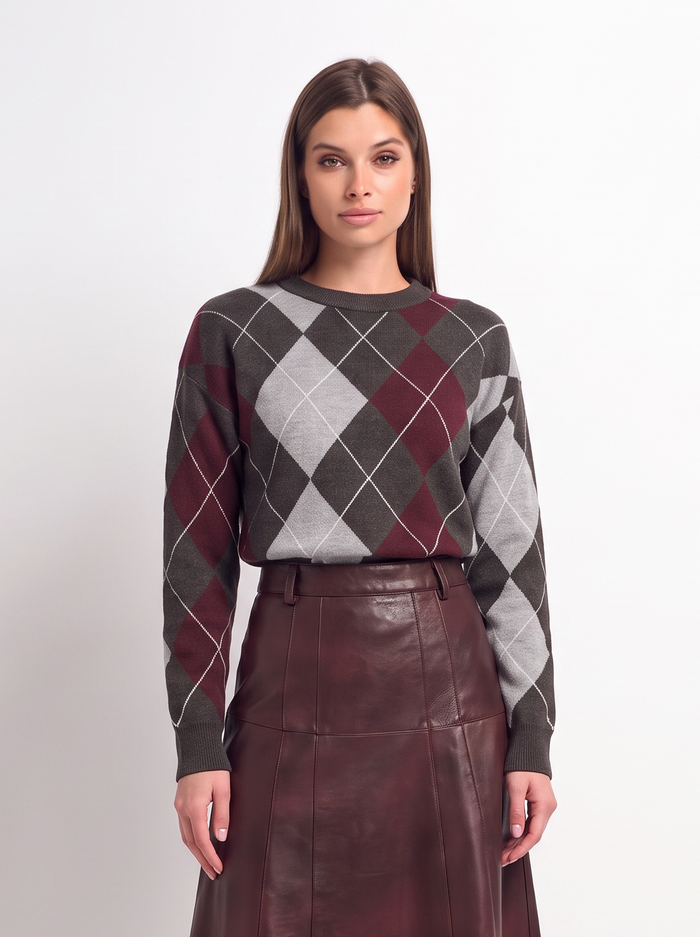 A woman stands against a plain light gray studio background wearing a long-sleeve argyle sweater in dark gray, burgundy, and white, paired with a dark burgundy pleated midi skirt and black pointed heels.