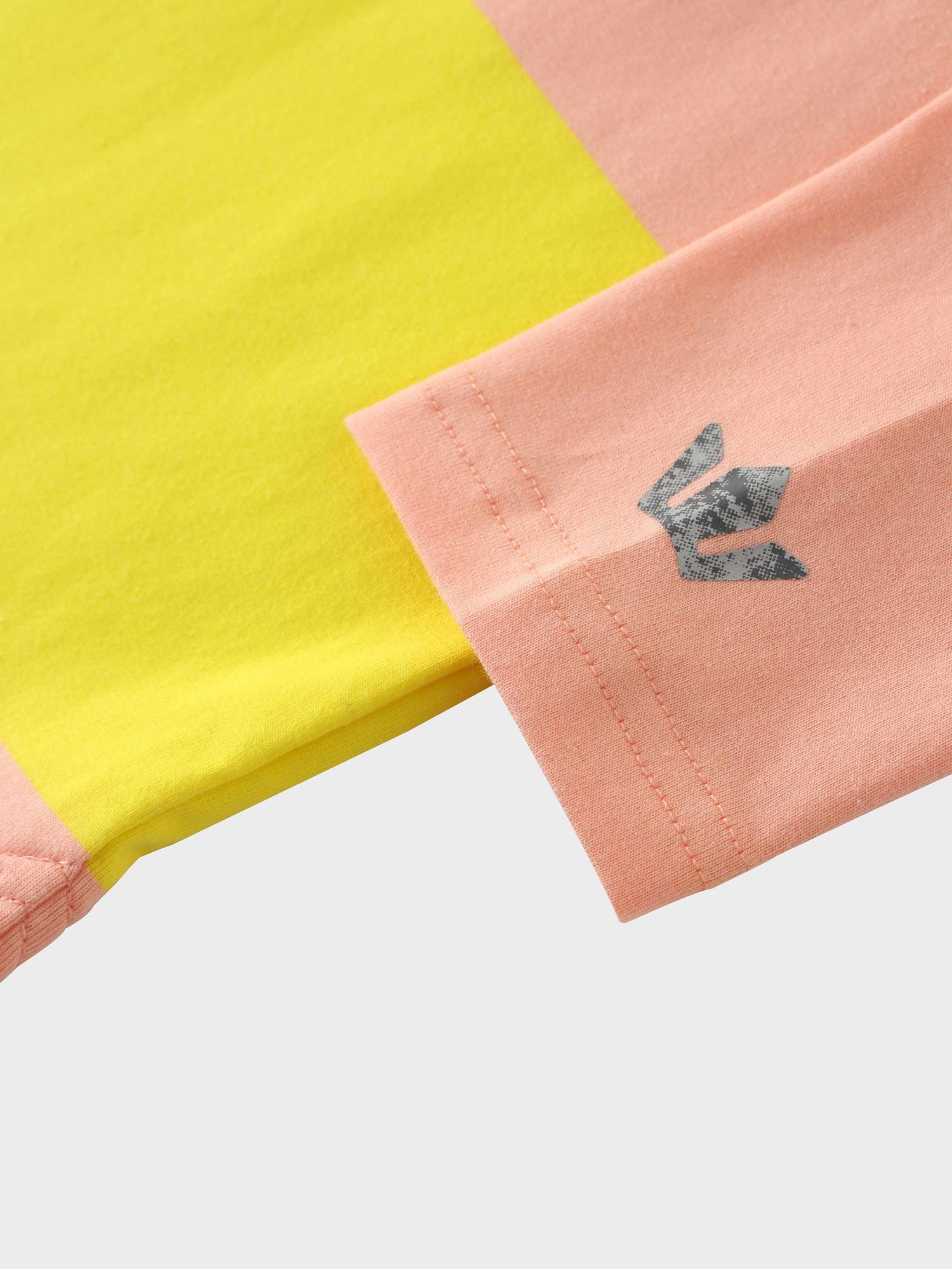 A close-up of the sleeve on the color-blocked shirt. The cuff is made of pink fabric and has a dark gray, textured logo printed on it, seen here next to the yellow fabric of the shirt&