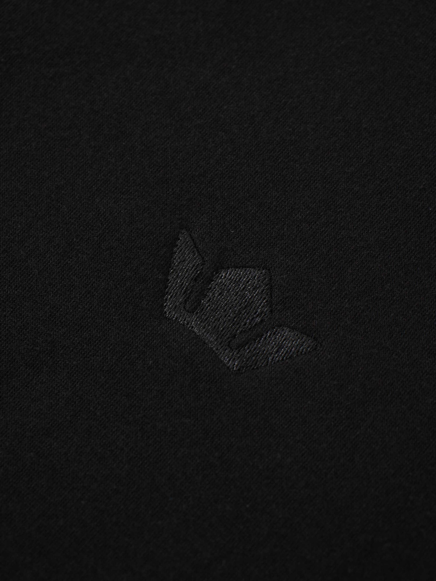 A macro shot of the black fabric, featuring a small, embroidered logo. The logo is stitched with matching black thread, creating a subtle, black-on-black effect.