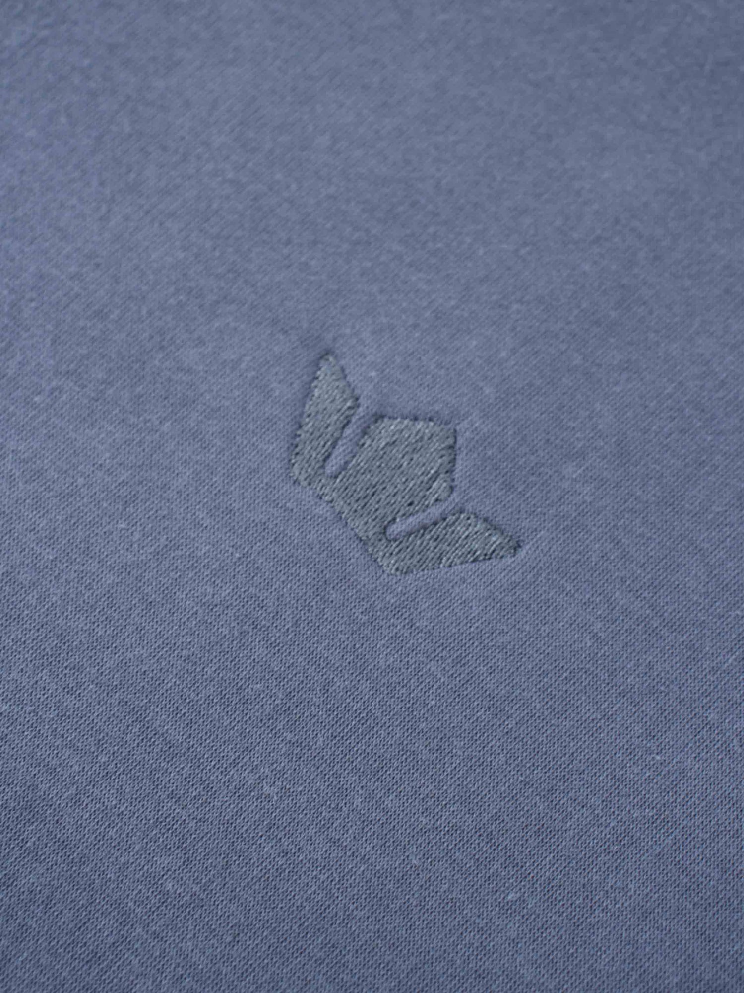 A macro shot of the dusty-blue fabric, showing a small, embroidered logo. The logo is stitched in a matching blue thread, creating a subtle, tone-on-tone effect.