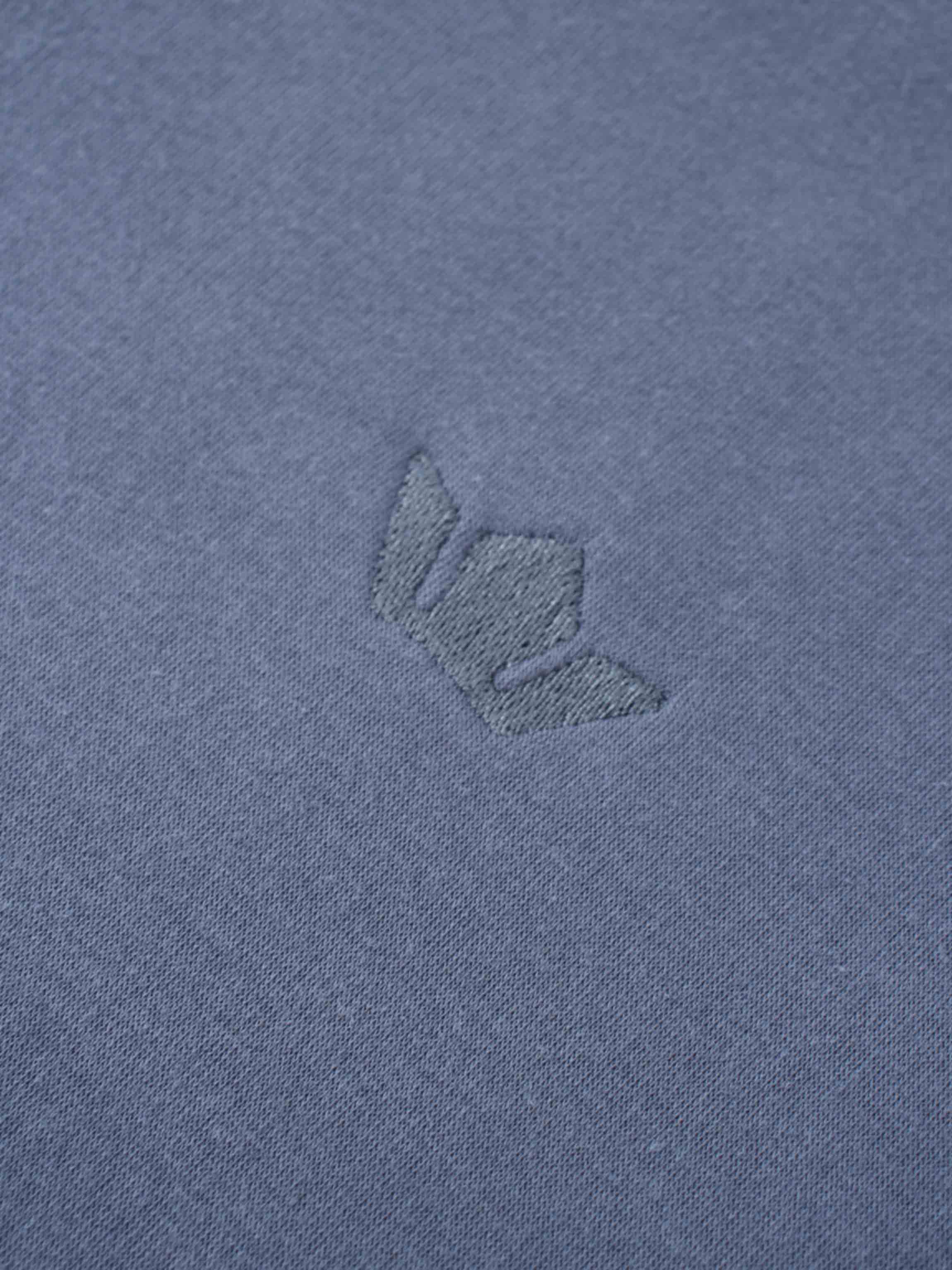 A macro shot of the dusty-blue fabric, showing a small, embroidered logo. The logo is stitched in a matching blue thread, creating a subtle, tone-on-tone effect.
