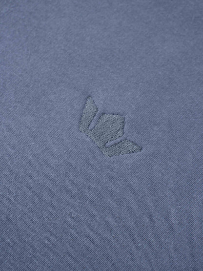 A macro shot of the dusty-blue fabric, showing a small, embroidered logo. The logo is stitched in a matching blue thread, creating a subtle, tone-on-tone effect.