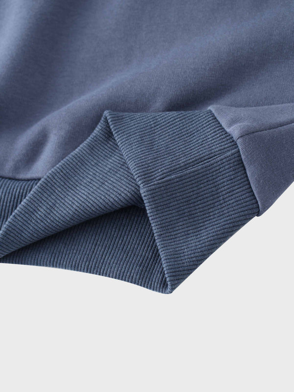 A full, front view of the dusty-blue sweatshirt. It has a ribbed crewneck collar, cuffs, and bottom hem, with the small, tone-on-tone embroidered logo visible on the left chest.