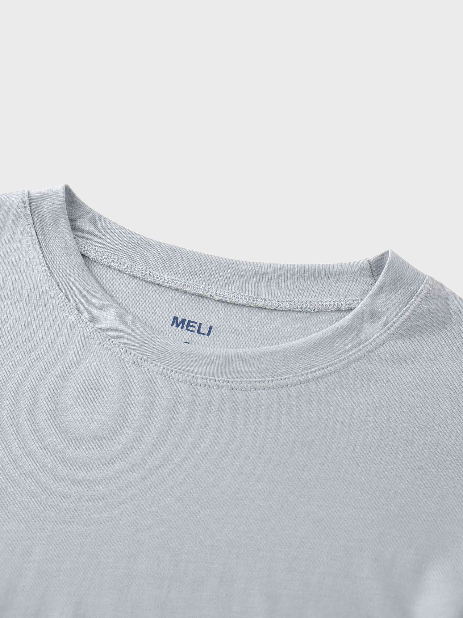 A close-up of the crewneck collar of the light gray t-shirt. On the inside, the brand name &quot;MELI&quot; is printed directly onto the fabric in blue.
